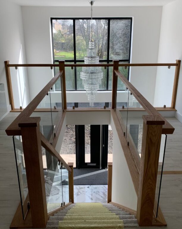 Elegant oak staircase with glass balustrade overlooking hallway with crystal chandelier by Overgreen Joinery