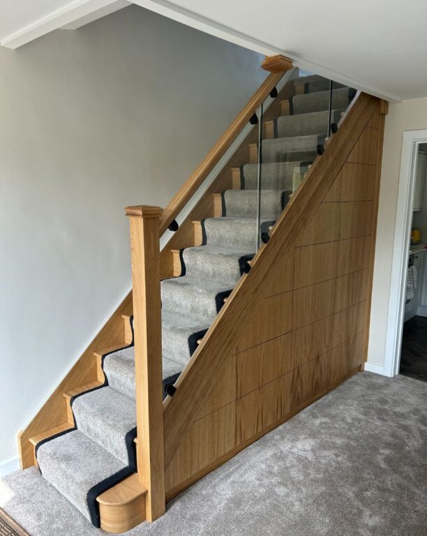 Oak staircase with glass balustrade and decorative geometric side panels with grey carpet runner by Overgreen Joinery