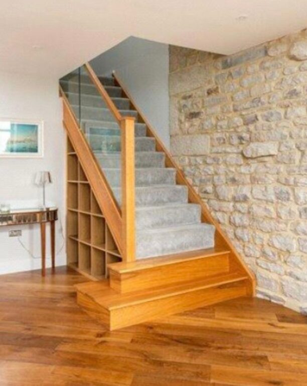 Oak staircase with glass balustrade and built-in storage against exposed stone wall by Overgreen Joinery